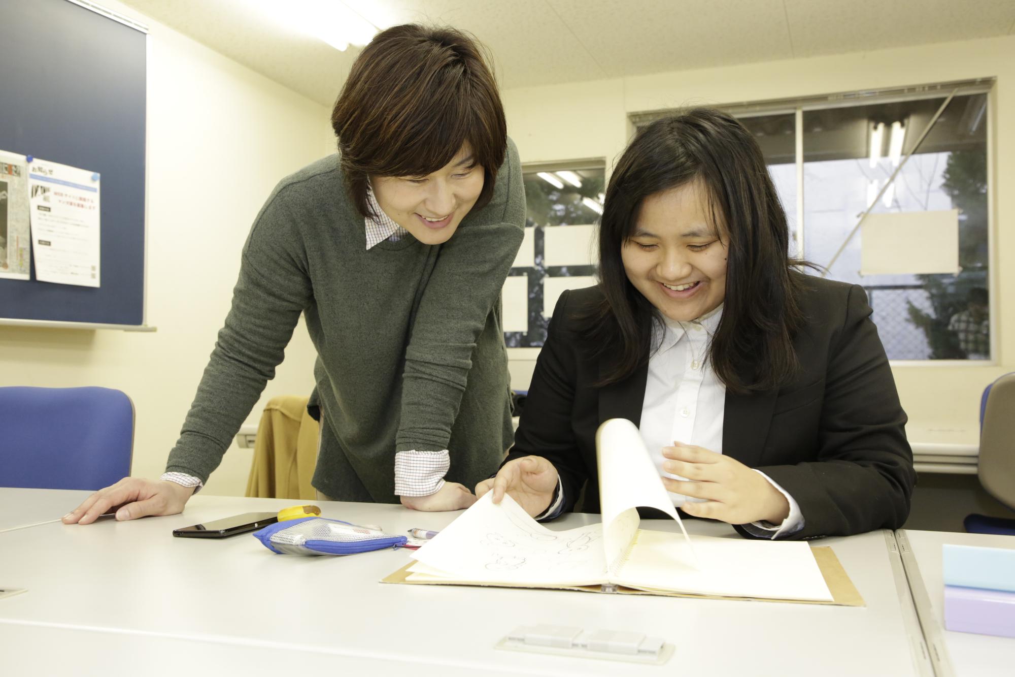 西宮市こども未来センターのパンフレット作成のための打ち合わせの様子を写した写真。デザイン案が描かれたスケッチブックを用いて学生が講師に説明している様子が写されている。