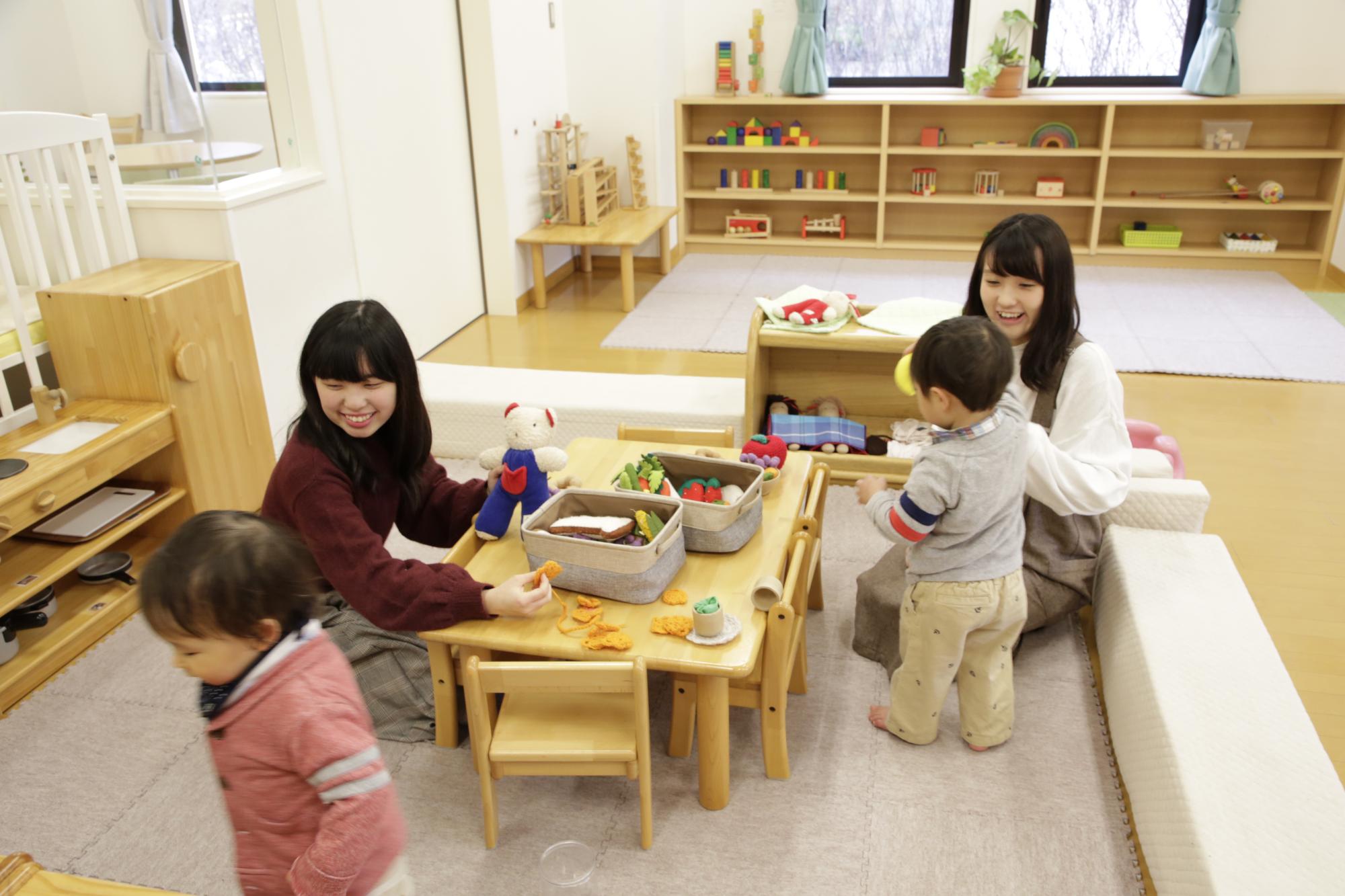 子供センターで行われてる子育て支援「さぽさぽ」で子供たちとふれあっている学生の様子を写した写真。中央に子供用の小さなテーブルや椅子が置かれており、ぬいぐるみ等のおもちゃが並んでいる。
