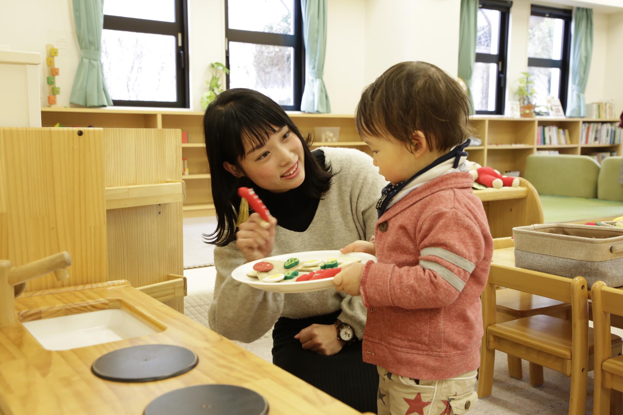 子供センターの取り組み「さぽさぽ」で子供と一緒に遊んでいる大学生ボランティアの姿が写っている写真。キッチンを模した木製のおもちゃが置かれている。